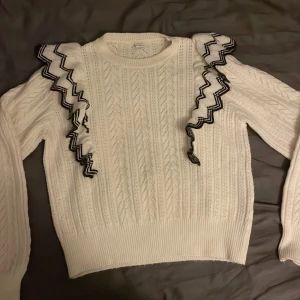 Vit stickad tröja med volangdetaljer - Säljer en vit stickad tröja med snygga svarta volangdetaljer längs axlarna. Tröjan har ett fint mönster och långa ärmar, perfekt för kyligare dagar. Passar både till jeans och kjol!