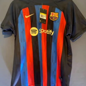 FC Barcelona fotbollströja från Nike - Snygg FC Barcelona tröja med klassiska rödblå ränder och sponsorloggor från Spotify och UNHCR. Tröjan är kortärmad och har klubbmärket på bröstet. Perfekt för alla Barça-fans! Shorts medföljer 