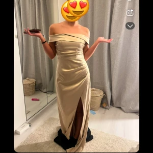 Champagnefärgad axelbandslös klänning - Elegant champagnefärgad långklänning med offshoulder-design och en stilren slits framtill. Perfekt för festliga tillfällen.