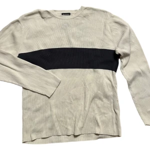 Beige och svart ribbad tröja från Polo Jeans Co. Ralph Lauren - Säljer en stilren beige tröja från Polo Jeans Co. Ralph Lauren med en bred svart rand över bröstet. Tröjan är ribbad och har långa ärmar, perfekt för en avslappnad look.