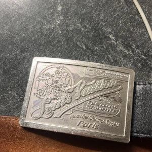 Vinted skärp med silverdetaljer från Louis Vuitton - Snyggt svart skärp i skinn från Louis Vuitton med en stor, rektangulär spänne i silver. Spännet har en detaljerad gravyr med texten 'Louis Vuitton Paris'. Perfekt för att ge en lyxig touch till din outfit.