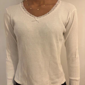 Vit långärmad topp med spetsdetaljer - BRANDY MELVILLE Säljer en stilren vit långärmad topp med v-ringning och spetsdetaljer runt halsen. Toppen har en liten rosett framtill och är tillverkad i ett ribbat material som ger en skön passform.