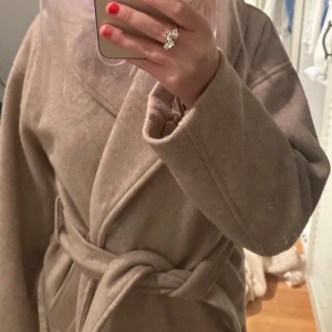 Beige kappa med knytband - Säljer en stilren beige kappa med knytband i midjan. Den har en omlott-design och långa ärmar, perfekt för kyligare dagar. Kappan är både elegant och bekväm, vilket gör den till ett mångsidigt tillskott i garderoben.