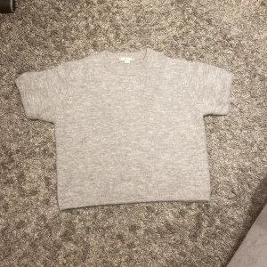 Grå stickad tröja från Gina Tricot - Mysig grå stickad tröja från Gina Tricot. Perfekt för kyliga dagar med sin mjuka och bekväma passform. Tröjan har korta ärmar och en klassisk rund halsringning.