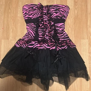 Zebramönstrad klänning - Snygg axelbandslös kort klänning med rosa och svart zebramönster. Den har en volangdetalj, sidenbands rosetter och två snörningar framtill och en baktill. Som korsett. Tyll och spets nedtill på klänningen. Ej använd och prislappen är kvar. Storlek L-XL, men går att dra åt med snörerna. Finns axelband att ta på. Nypris 500kr💗