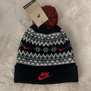 Svart och vit mössa från Nike - Snygg svart och vit mössa från Nike med rött broderat Nike-märke och mönster. Mössan har en stor tofs i rött och svart på toppen och ett klassiskt vintermönster med snöflingor och sicksackmönster. Hör av er vid funderingar och pris kan diskuteras vid snabb affär mvh Malte!