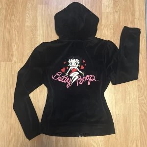 Svart tröja med Betty Boop - Snygg svart velourtröja med huva och dragkedja. På baksidan finns ett stort Betty Boop-motiv med röda hjärtan. Dragkedjan har en hjärtformad dragkedjekläpp med Betty Boop-design. Storlek S. Mycket bra skick då den knappt är använd💗