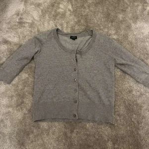 Grå kofta från Lindex - Säljer en stilren grå kofta från Lindex med knappar framtill. Koftan har trekvartsärmar och ribbade kanter vid ärmslut och nederkant. 