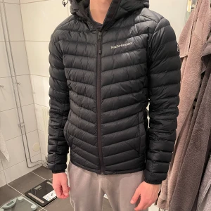 Marinblå dunjacka från Peak Performance - Snygg och stilren svart dunjacka från Peak Performance. Jackan har en dragkedja framtill och är långärmad med en huva för extra värme. Perfekt för kyliga dagar. Sparsamt använd. 