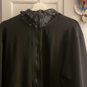 Louis Vuitton tracksuit - Snygg svart jacka med dragkedja och monogrammönstrad huva. Perfekt för en stilren look. Jackan har långa ärmar och en modern design. Säg till om ni vill ha bild på byxorna!! Pris ändras om man ska ha med båda