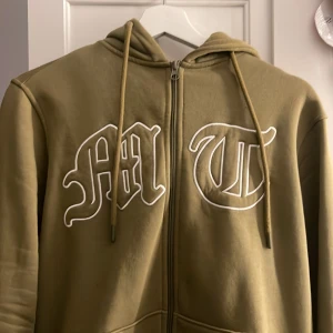 Tuttolente tröja  - Säljer en olivgrön hoodie med dragkedja och tryck i vitt på bröstet. Den har en justerbar huva med snören och en bekväm passform. Perfekt för en avslappnad stil.