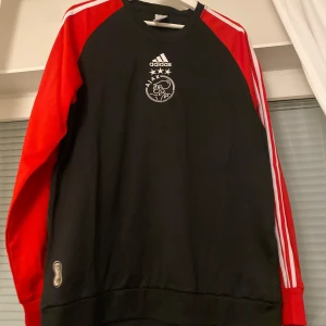 Ajax jumper från Adidas - Snygg svart och röd tröja från Adidas med Ajax-logga på bröstet. Tröjan har långa ärmar med vita ränder längs sidorna och en stor Adidas-logga på ryggen. Perfekt för fotbollsfans!