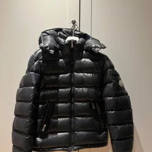 Svart dunjacka från Moncler - Snygg svart dunjacka från Moncler med huva och dragkedja. Jackan har en glansig finish och Moncler-logga på ärmen. Perfekt för kyliga dagar.Priset kan diskuteras vid snabb affär. 