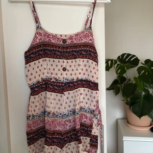 Mönstrad sommarbyxdress med knappar - Säljer en mönstrad sommarbyxdress med smala axelband och knappar framtill. Byxdressen har ett färgglatt mönster i rött, blått och svart på en beige bakgrund. Perfekt för varma dagar!