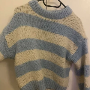 Randig stickad tröja i blått och vitt - Mysig stickad tröja med breda ränder i ljusblått och vitt. Tröjan har en avslappnad passform och långa ärmar, perfekt för kyliga dagar. Den ribbade halsringningen och ärmsluten ger en extra touch av komfort.