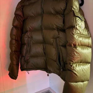 Olivgrön dunjacka från Moncler - Säljer en snygg olivgrön dunjacka från Moncler. Jackan har en quiltad design med en praktisk dragkedja och en ficka med knappdetalj på ärmen. Perfekt för kyliga dagar!