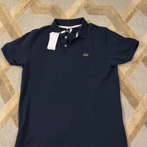 Mörkblå pikétröja från Lacoste - Snygg mörkblå pikétröja från Lacoste med klassisk krage och knappar framtill. Tröjan har korta ärmar och det ikoniska krokodilmärket på bröstet. Perfekt för en stilren look.