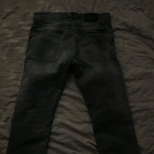 Grå jeans från Nudie Jeans - Snygga grå jeans från Nudie Jeans med klassisk femficksdesign och subtila slitningar. De har en dragkedja och knapp framtill samt en broderad detalj på bakfickan. Perfekta för en avslappnad stil. W29 L 32 pris ej hugget i sten