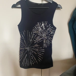 Svart ärmlös topp med glittrande paljetter från Hugo Boss - Snygg svart ärmlös topp från Hugo Boss med glittrande paljettdetaljer i ett fyrverkeri liknande design. Toppen har en elegant och stilren design. Jag skulle säga att den är S/M i storlek. Måste handtvätts. Använd: Aldrig. Orsak för säljning: Använder ej!❣️