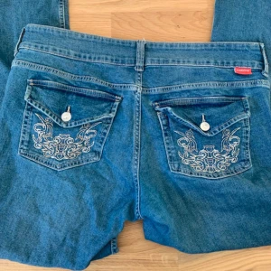 Blå jeans med broderade bakfickor - Slutsålda HM bootcut jeans. Snygga blå jeans med unika broderade detaljer på bakfickorna. De har en klassisk femficksdesign och knappar framtill. Perfekta för en avslappnad stil med en touch av elegans. Använda max 5 gånger, top skick! Skriv om du undrar nåt!😊🔥