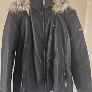 Svart jacka med fuskpäls från Tommy Jeans - Snygg svart jacka från Tommy Jeans med fuskpälsdetaljer på huvan. Jackan har dragkedja framtill och en liten logotyp på ärmen. Perfekt för kyligare dagar.