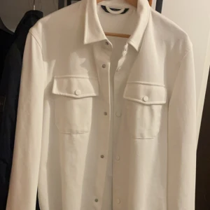 Vit overshirt med knappar - Snygg vit overshirt med knappar framtill och två bröstfickor med lock. Perfekt för lager-på-lager-stil och passar till många olika outfits. Enkel och stilren design som funkar året runt.