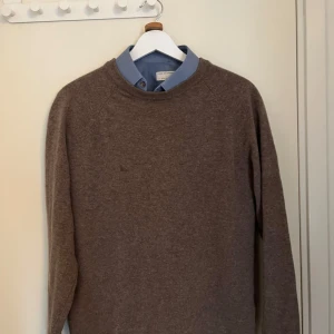 Brun tröja från Les Deux - Säljer en stilren brun tröja från Les Deux. Tröjan har långa ärmar och en liten detalj i form av en broderad flagga på ärmen. Perfekt för en avslappnad och snygg look.