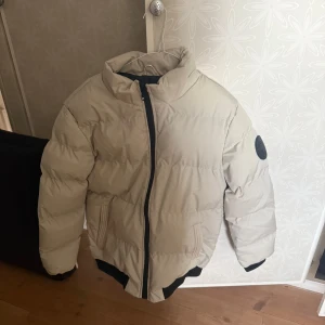 Grå pufferjacka med svart emblem - Snygg grå pufferjacka med svart dragkedja och ett coolt svart emblem på ärmen. Jackan har en hög krage och elastiska muddar vid ärmslut och nederkant för extra värme. Perfekt för kyliga dagar! Helt ny aldrig använd 