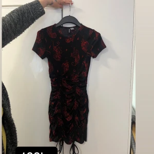 Svart och röd klänning med mönster - Snygg svart klänning med röda mönster och korta ärmar. Den har en åtsittande passform och snörning framtill som ger en cool look. Perfekt för en kväll ute eller en speciell tillställning.