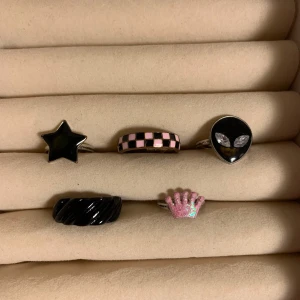 Fem unika ringar i olika stilar - Säljer ett set med fem coola ringar. En svart stjärnring, en rosa och svart schackmönstrad ring, en svart ring med alienmotiv, en svart vriden ring och en rosa glittrig kronring. Perfekt för att ge din stil en unik touch! (15kr för alla ringar)
