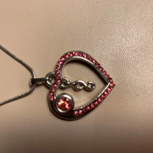 Halsband med hjärthänge och rosa stenar - Elegant halsband med ett hjärthänge dekorerat med små rosa stenar och en större rosa sten i mitten. Hänget har en unik design med en oändlighetssymbol inuti hjärtat. Kedjan är tunn och silverfärgad med justerbar längd. Använd 1 gång.