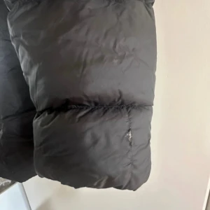 Svart dunjacka från Zara - Snygg svart dunjacka från Zara med pälskantad huva och dragkedja framtill. Jackan har en figursydd passform och är perfekt för kyliga dagar. Den har långa ärmar och en stilren design.