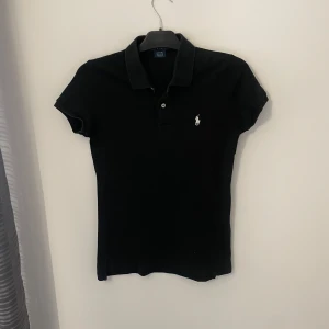 Svart pikétröja från Ralph Lauren - Säljer en klassisk svart pikétröja från Ralph Lauren med korta ärmar och den ikoniska loggan broderad på bröstet. Tillverkad i 100% bomull för en bekväm passform. Perfekt för en stilren look.