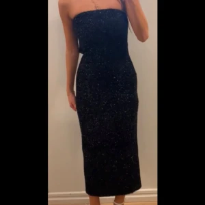 Svart glittrig axelbandslös klänning - Snygg svart axelbandslös klänning med glittrigt material som ger en festlig look. Klänningen har en tight passform och en slits baktill för extra rörelsefrihet. Perfekt för en kväll ute eller en speciell tillställning. Klänningen är endast använd en gång🥰