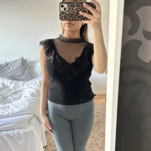 Svart topp med spetsdetaljer - Snygg svart topp med spetsdetaljer och transparenta partier vid halsen. Toppen har en vacker volangkant och är ärmlös, vilket ger en elegant look. Perfekt för att piffa upp vilken outfit som helst.