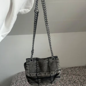 Grå axelväska med kedja - Snygg grå axelväska med kedjedetaljer och ett coolt rutigt mönster. Väskan har en praktisk dragkedja och en knappstängning. Perfekt för att ge din outfit en edgy touch.