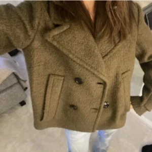 Olivgrön kappa - Säljer en stilren olivgrön kappa med dubbelknäppning och breda slag. Jackan har en klassisk design med långa ärmar och två framfickor. Perfekt för kyligare dagar.