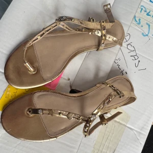 Guldiga sandaler från Aldo - Snygga guldiga sandaler från Aldo med dekorativa stjärnor och pärlor på remmarna. Sandalerna har en justerbar rem runt ankeln och en platt sula för bekväm passform. Perfekta för att ge en glittrig touch till din outfit.