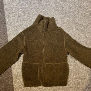 Grön teddyjacka med dragkedja - Mysig gröna teddyjacka från H&M med dragkedja framtill. Jackan har en hög krage och två stora fickor. Perfekt för kyliga dagar när du vill hålla dig varm och bekväm.