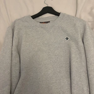 Grå sweatshirt från Morris - Säljer en stilren grå sweatshirt från Morris med ett litet emblem på bröstet. Tröjan har en klassisk rund halsringning och långa ärmar. Perfekt för en avslappnad look.