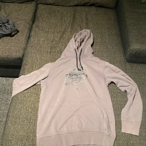 Lager 157 - Säljer en beige hoodie med tryck på framsidan. Den har en klassisk design med huva och långa ärmar. Perfekt för en avslappnad stil.