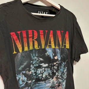 Svart Nirvana t-shirt från Stay/Carlings - Cool svart t-shirt från Stay/Carlings med stort Nirvana-tryck i rött och gult på framsidan. T-shirten har korta ärmar och en rund halsringning. Perfekt för alla Nirvana-fans eller den som vill ha en avslappnad stil.