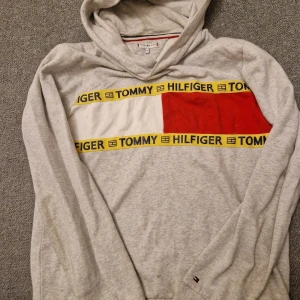 Grå hoodie från Tommy Hilfiger - Säljer en stilren grå hoodie från Tommy Hilfiger med ett iögonfallande rött och vitt band över bröstet med märkets namn. Tröjan har långa ärmar och en klassisk huva. Perfekt för en avslappnad stil. 🧢
