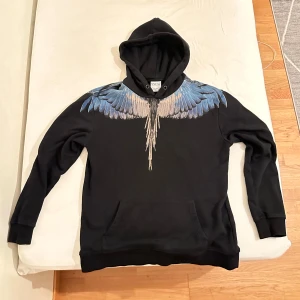 Svart hoodie med blå vingmönster - Säljer en svart hoodie från Marcelo Burlon med ett coolt blått vingmönster som sträcker sig över axlarna och ryggen. Tröjan har en klassisk passform med långa ärmar och en praktisk magficka. Perfekt för dig som vill sticka ut med stil! 🖤💙