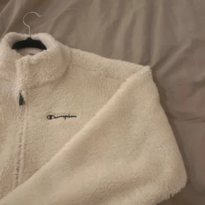 Vit teddyjacka från Champion - Mysig vit teddyjacka från Champion med dragkedja framtill. Perfekt för kyliga dagar med sin fluffiga och varma design. Jackan har en broderad logga på bröstet och långa ärmar.