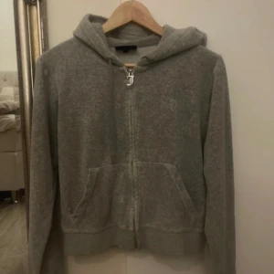 Grå hoodie från Juicy Couture - Säljer en grå hoodie från Juicy Couture med dragkedja och huva. Den har en broderad logga på bröstet och en snygg dragkedja med ett 'J'-hänge. Perfekt för en avslappnad stil.