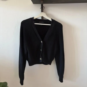 Svart stickad kofta med knappar - Säljer en stilren svart stickad kofta med tre knappar framtill. Koftan har en v-ringad design och långa ärmar, perfekt för kyligare dagar. Den är både bekväm och lätt att matcha med olika outfits.