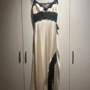 Champagnefärgad långklänning med spetsdetaljer - Elegant champagnefärgad långklänning med svarta spetsdetaljer. Klänningen har smala axelband och en slits på sidan. Perfekt för en glamorös kväll. Storlek 42 men skulle säga att den är liten i storleken