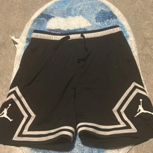 Svarta shorts med grå detaljer från Jordan - Snygga svarta shorts från Jordan med grå detaljer och det ikoniska logotyptrycket på benen. De har en elastisk midja med snörning för justerbar passform. Perfekta för sport och avslappnad stil.