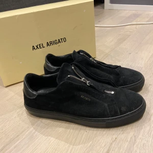 Svarta sneakers från Axel Arigato - Snygga svarta sneakers i mocka från Axel Arigato med dragkedja framtill. Perfekta för en stilren look. Skorna har en låg profil och en diskret logga på sidan perfekta till våren och sommaren.